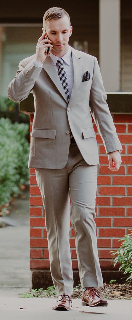 A man walking outdoors in a well-fitted light tan suit with a checked tie and pocket square