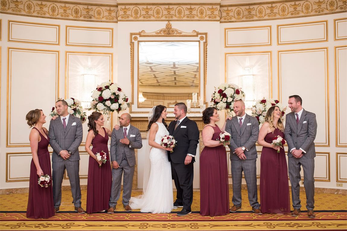 A wedding party in an ornate ballroom, the groom in a dark charcoal suit set apart from his groomsmen in grey suits with burgundy ties